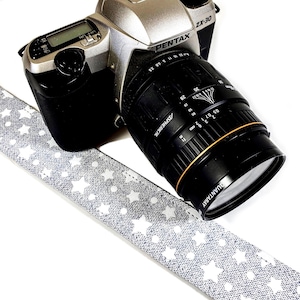 May include: A silver Pentax ZX-30 camera with a black lens attached. The camera is attached to a gray and white camera strap with a star pattern.