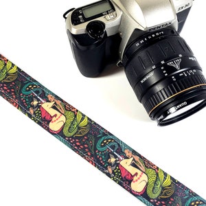 Peut inclure: Un sangle d'appareil photo noir et vert avec un motif coloré de sirène et de licorne. La sangle présente un motif répété de sirènes et de licornes avec des accents floraux.