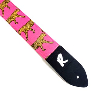 May include: Pink guitar strap with a leopard print design and a black leather end with the letter "R" in white.