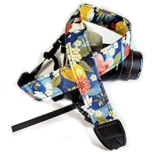 May include: A camera strap with a floral pattern in shades of blue, red, yellow, and white. The strap is wrapped around a camera with a black lens. The strap has black plastic buckles and leather ends.