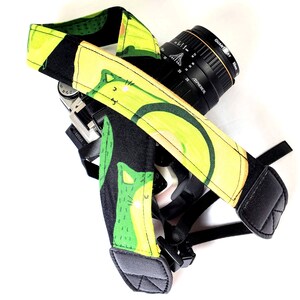 May include: A black and green camera strap with a pattern of avocados and cats. The strap is attached to a black camera lens with the text "QUANTIBAY" and "ASPHERIC" printed on it.