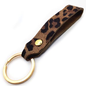 May include: A keychain with a leopard print strap and a gold-colored key ring. The strap is brown with black spots and is secured with a gold-colored button. The key ring is a large, circular design.