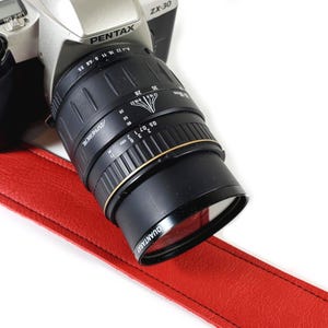 May include: A silver Pentax ZX-30 camera with a black lens and a red leather strap. The lens has the text "QUANTARAY" and measurement markings. The camera is on a white background.