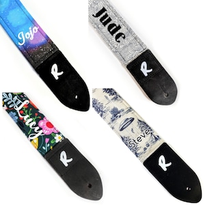 May include: Four guitar straps with different designs and personalized names. The straps are black with white lettering and have a white "R" logo. The designs include a galaxy print with the name "Jojo", a wave print with the name "Jude", a floral print with the name "Lucy", and a UFO print with the name "Steve".