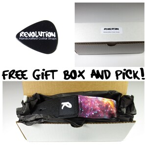 May include: A black guitar pick with the word "REVOLUTION" and "Handcrafted Guitar Straps" in white. A guitar strap with a galaxy design is in a gift box. The text "FREE GIFT BOX AND PICK!" is also visible.