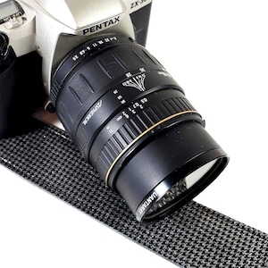 May include: A silver Pentax ZX-30 camera with a black lens. The lens has a gold ring and the text "QUANTARAY" is visible. The camera strap is black and white houndstooth. The lens has measurement markings.