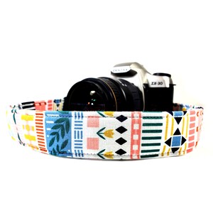 May include: A white camera strap with a colorful geometric pattern. The strap features a variety of colors, including blue, green, yellow, pink, and orange. The strap is designed to be used with a camera, and it is a great way to add a touch of personality to your photography gear.
