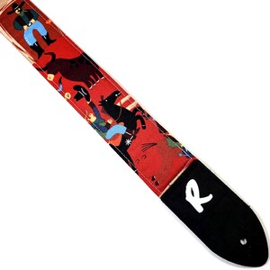 May include: Red guitar strap with a cowboy-themed pattern. The strap features illustrations of cowboys, horses, and a bull. The end of the strap is black with a white "R" logo.