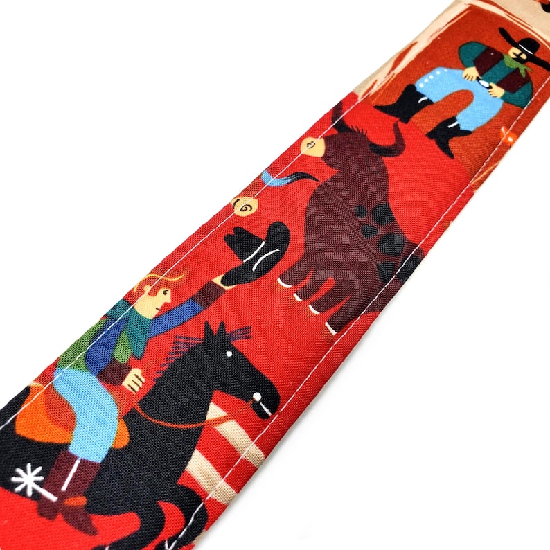 May include: Red fabric with a cowboy-themed pattern. The design features cowboys, a bull, and horses in a stylized illustration. The fabric is likely used for accessories or crafts.