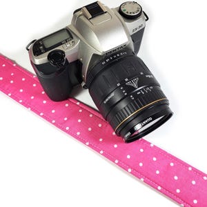 Op de afbeelding: Een zilveren en zwarte Pentax ZX-30 camera met een zwarte lens en een roze polka dot camerariem. De riem is felroze met witte stippen. De cameralens heeft een Quantaray 55mm filter.