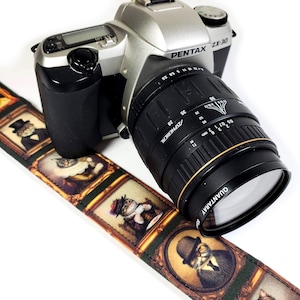 May include: A brown camera strap with a repeating pattern of gold frames containing images of cats wearing hats and clothing. The strap is attached to a silver Pentax ZX-30 camera with a black lens. The lens has the text "QUANTARAY" and "ASPHERICAL" printed on it.