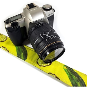 以下が含まれることがあります： 黒いレンズと黄色のカメラストラップが付いた、シルバーとブラックのPentax ZX-30カメラ。ストラップには緑色のキュウリの模様があります。カメラは白い背景にあります。