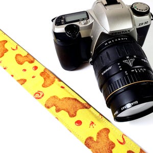 May include: A yellow camera strap with a pattern of fried chicken and red sauce. The strap is attached to a silver Pentax ZX-30 camera with a black lens.