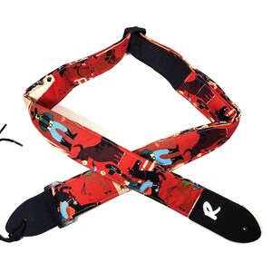 May include: A red guitar strap with a repeating pattern of cowboys, dogs, and other figures. The strap has black leather ends and a black adjustable section. The strap is designed to be worn over the shoulder while playing a guitar.