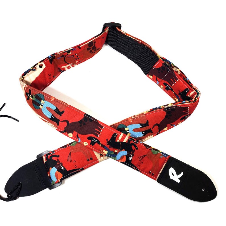 May include: A red guitar strap with a repeating pattern of cowboys, dogs, and other figures. The strap has black leather ends and a black adjustable section. The strap is designed to be worn over the shoulder while playing a guitar.