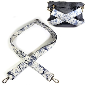 May include: A dark grey crossbody bag with a detachable strap. The strap is cream-coloured with a repeating blue octopus design. The strap has gold-coloured metal clasps for attachment. The bag's strap is crossed over the bag.