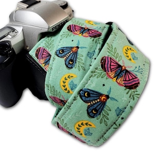 May include: A camera strap with a teal background featuring a repeating pattern of moths, crescent moons, and leaves. The moths have pink, yellow, and blue wings. The strap is attached to a silver Pentax camera.