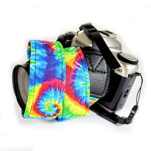 May include: A silver Pentax camera with a black leather strap and a colourful tie-dye camera strap. The tie-dye strap features a vibrant mix of blue, red, yellow, and green hues. The camera lens is visible.