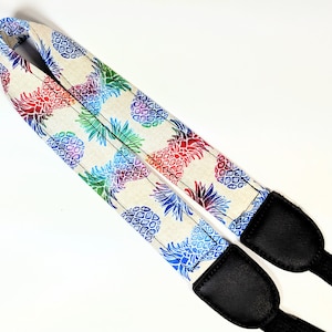 Könnte beinhalten: Ein Kameragurt mit weißem Hintergrund und einem bunten Ananas-Muster. Der Gurt ist aus Stoff und hat schwarze Lederenden.