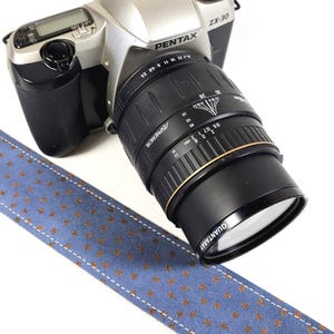 May include: A silver Pentax ZX-30 camera with a black lens attached. The lens has a gold ring with the text "QUANTARAY" and "ASPHERICAL" printed on it. The camera is attached to a blue camera strap with a pattern of small gold stars.