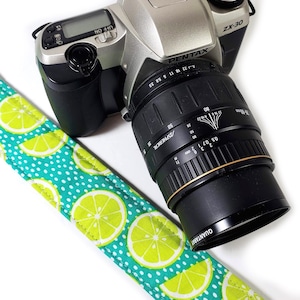 May include: A teal and white camera strap with a lime green lemon print. The strap is attached to a silver Pentax ZX-30 camera with a black lens.