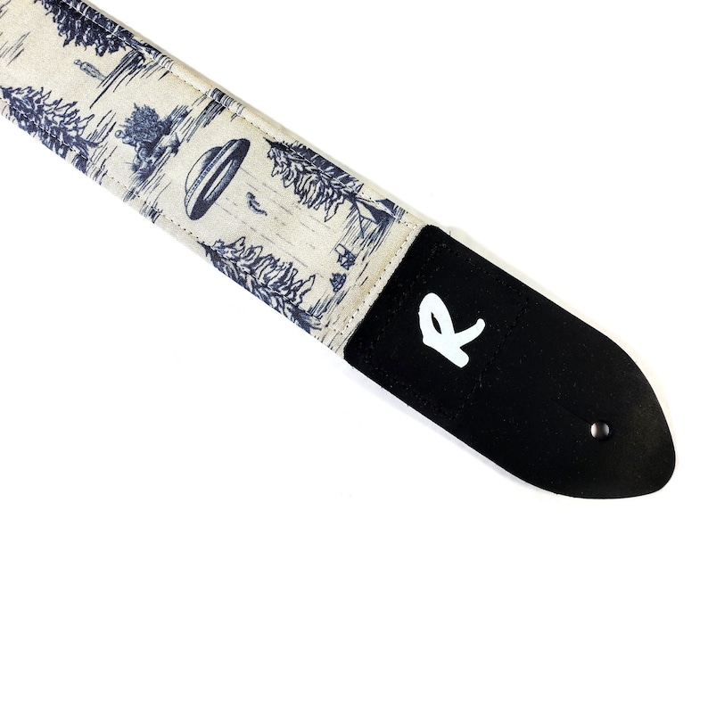 Pode incluir: Uma correia de guitarra branca com uma estampa de alien&iacute;gena e floresta azul e branca. A correia tem uma ponta de couro preto com um "R" branco impresso nela.