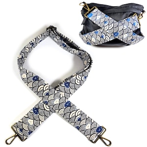 May include: A patterned bag strap with a repeating design of white, navy blue, and black wave-like shapes. The strap has gold-colored metal clasps on each end. A dark gray bag with a matching strap is also visible.