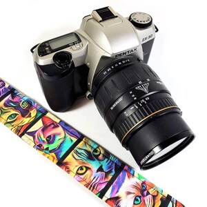 May include: A silver Pentax ZX-30 film camera with a black lens attached. The camera has a black and white cat-themed strap.