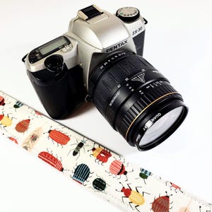 May include: A silver Pentax ZX-30 camera with a black lens and a white camera strap with a colorful bug pattern. The lens has the text "Vivitar Series 1" and "28-80mm 1:3.5-5.6" printed on it.