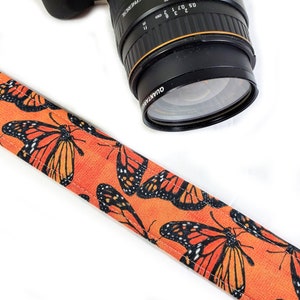 May include: Orange camera strap with a repeating pattern of black monarch butterflies. The strap is made of fabric and has a smooth texture.