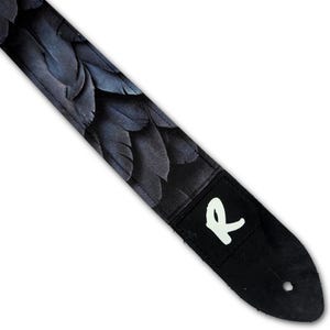 Peut inclure: Une sangle de guitare arborant un motif de plumes superposées dans des tons noirs et bleu profond. La sangle comprend une extrémité en cuir noir avec une lettre R blanche imprimée et un trou circulaire pour la fixation à un instrument.