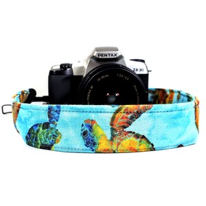 Könnte beinhalten: Ein blaues Kameraband mit einem Meeresschildkrötenmuster. Das Band ist an einer silbernen Pentax ZX-30 Kamera befestigt.