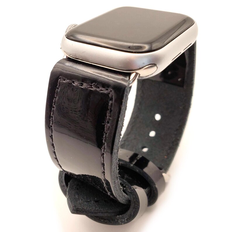 Puede incluir: Un Apple Watch plateado con una correa de charol negro. La correa tiene un acabado brillante y se asegura con una hebilla. La esfera del reloj es negra y la correa est&aacute; cosida con un hilo en contraste.