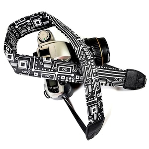 May include: A black camera strap with a white circuit board pattern. The strap is attached to a black camera with a silver lens.