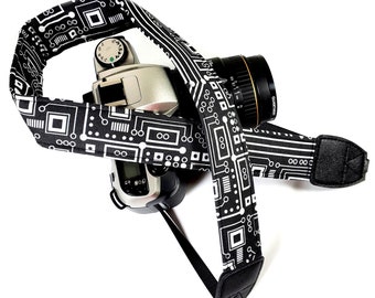 May include: A black camera strap with a white circuit board pattern. The strap is attached to a black camera with a silver lens.