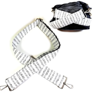 May include: White and black musical note patterned adjustable shoulder strap with silver hardware. The strap is shown lying flat and crossed over itself.