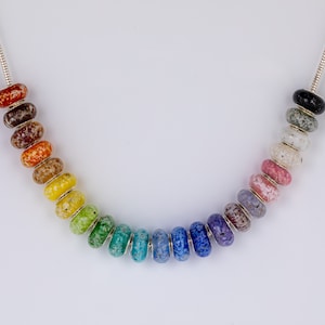 Op de afbeelding: Een zilveren ketting met een regenboog van gekleurde glazen kralen. Elke kraal heeft een gevlekt patroon.