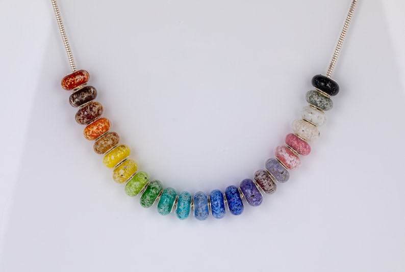 Op de afbeelding: Een zilveren ketting met een regenboog van gekleurde glazen kralen. Elke kraal heeft een gevlekt patroon.