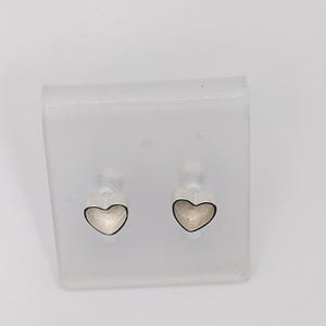 May include: A pair of heart-shaped stud earrings. Each earring features a silver-colored outline with a light-colored interior. The earrings are displayed on a clear, rectangular plastic holder against a white background.