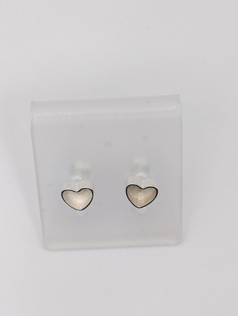 May include: A pair of heart-shaped stud earrings. Each earring features a silver-colored outline with a light-colored interior. The earrings are displayed on a clear, rectangular plastic holder against a white background.