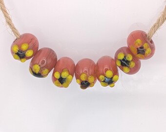 May include: A strand of handmade glass beads. The beads are a reddish-pink color with yellow and black floral designs. The beads are strung on a natural jute cord, and are displayed on a white surface.