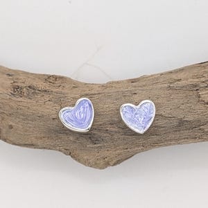 May include: A pair of heart-shaped stud earrings. Each earring features a silver border and a light purple enamel filling with a marbled effect. The earrings are displayed on a piece of driftwood.