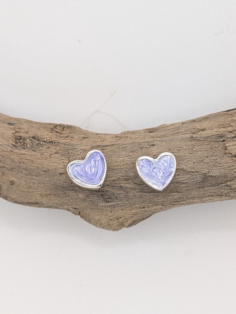 May include: A pair of heart-shaped stud earrings. Each earring features a silver border and a light purple enamel filling with a marbled effect. The earrings are displayed on a piece of driftwood.