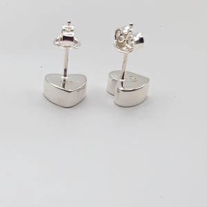May include: A pair of silver heart-shaped stud earrings. The earrings have a post and butterfly backing. The heart-shaped studs are smooth and polished, reflecting light. The earrings are set against a white background.