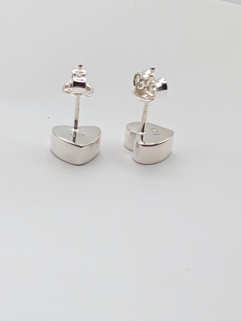 May include: A pair of silver heart-shaped stud earrings. The earrings have a post and butterfly backing. The heart-shaped studs are smooth and polished, reflecting light. The earrings are set against a white background.