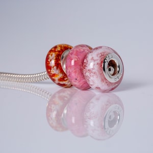 Op de afbeelding: Drie glazen kralen op een zilveren ketting. De kralen zijn rood, roze en wit met een marmereffect. De kraal rechts heeft een zilveren bedel met het nummer "925" erop.