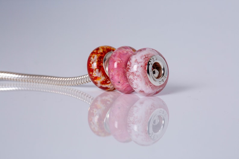 Op de afbeelding: Drie glazen kralen op een zilveren ketting. De kralen zijn rood, roze en wit met een marmereffect. De kraal rechts heeft een zilveren bedel met het nummer "925" erop.