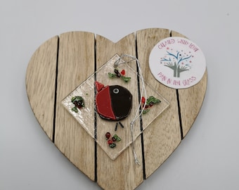 May include: A diamond-shaped clear glass ornament featuring a stylised red and black bird design with small green and red holly accents. A silver string is attached for hanging. A circular sticker reads Created With Love Pain In The Glass.