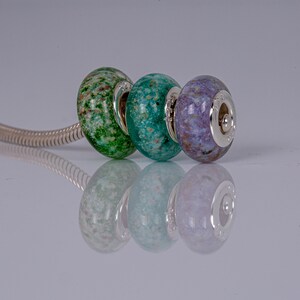 Op de afbeelding: Drie glazen kralen op een zilveren ketting. De kralen zijn groen, blauw en paars met een gevlekt patroon.