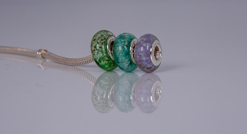 Op de afbeelding: Drie glazen kralen op een zilveren ketting. De kralen zijn groen, blauw en paars met een gevlekt patroon.
